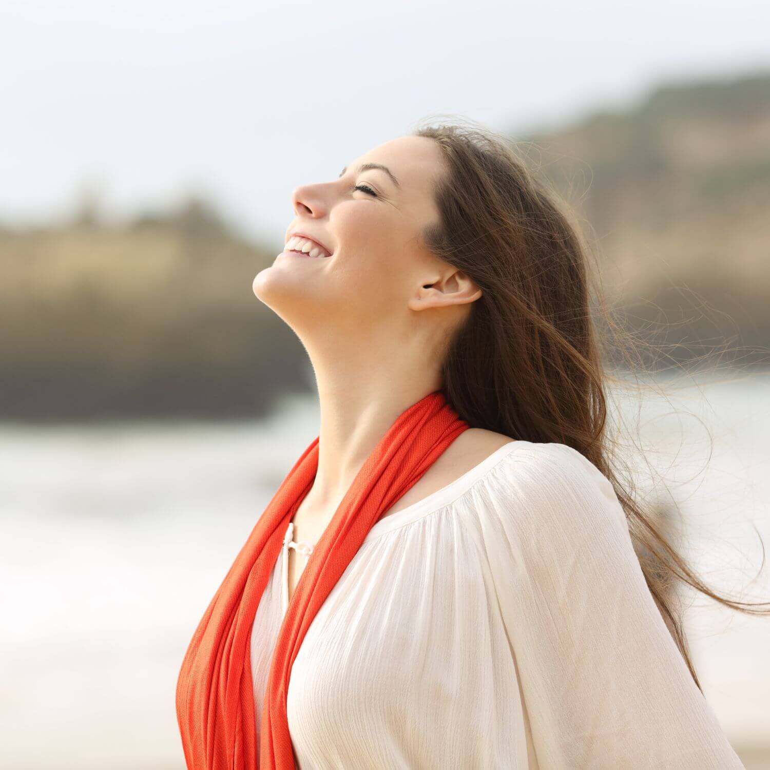 Lyxigt essentiellt hudplåster, kvinna med ett leende på stranden, bär en röd scarf.